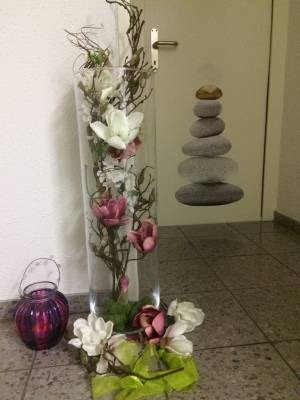 Eine hohe durchsichtige Vase mit einer Blume mit künstlichen Magnolienblüten. Die Vase enthält ausserdem trockenes Moos, Steine und Äste.